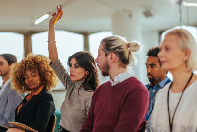 Curious,Young,Businesswoman,Raising,Hand,At,Multiracial,Group,Meeting,Engaging
